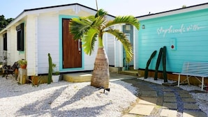 A building with a white exterior and a turquoise door, a palm tree in the center, and a bench to the right.