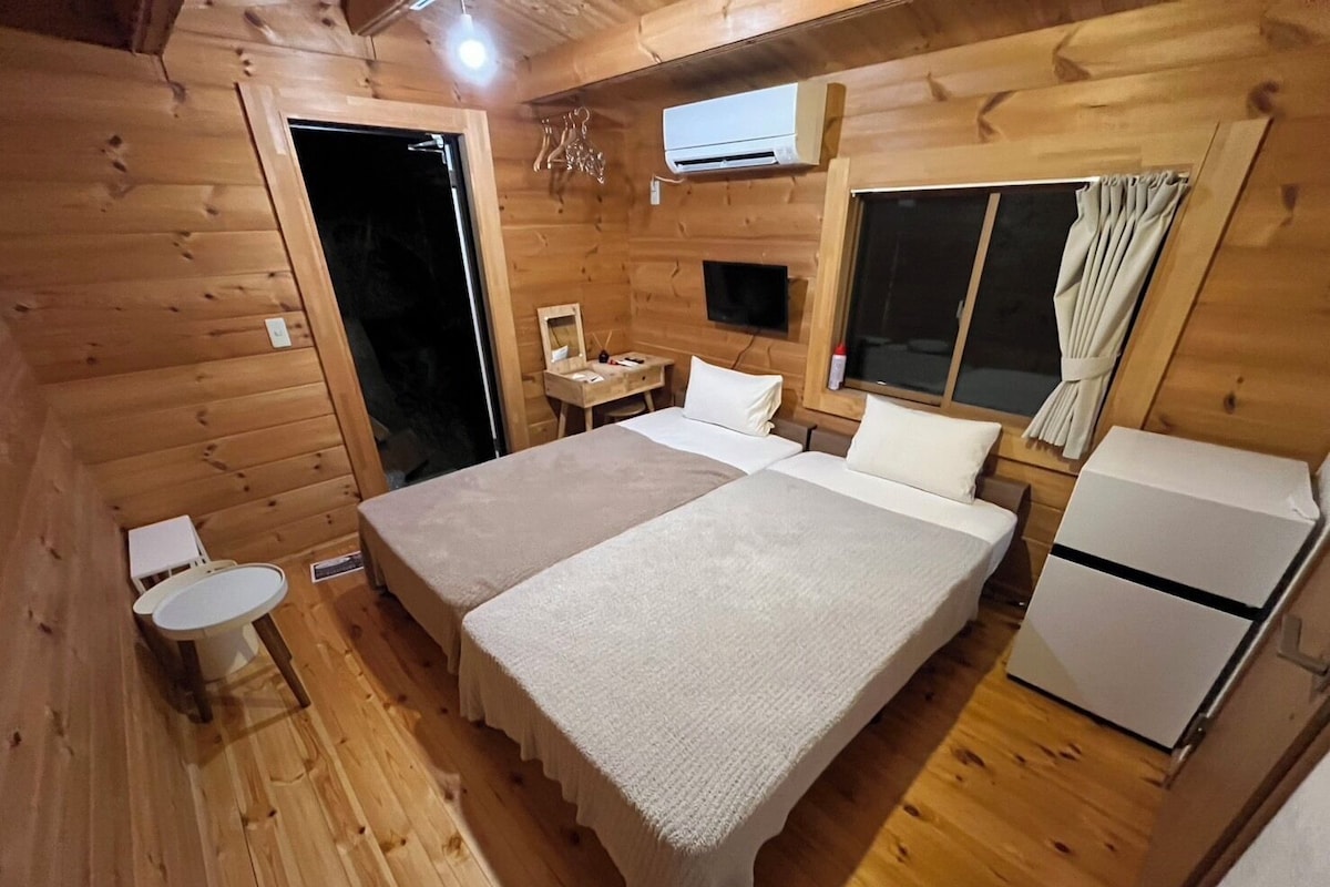 A wooden cabin room with two beds, a small table, a chair, a toilet, and a refrigerator.