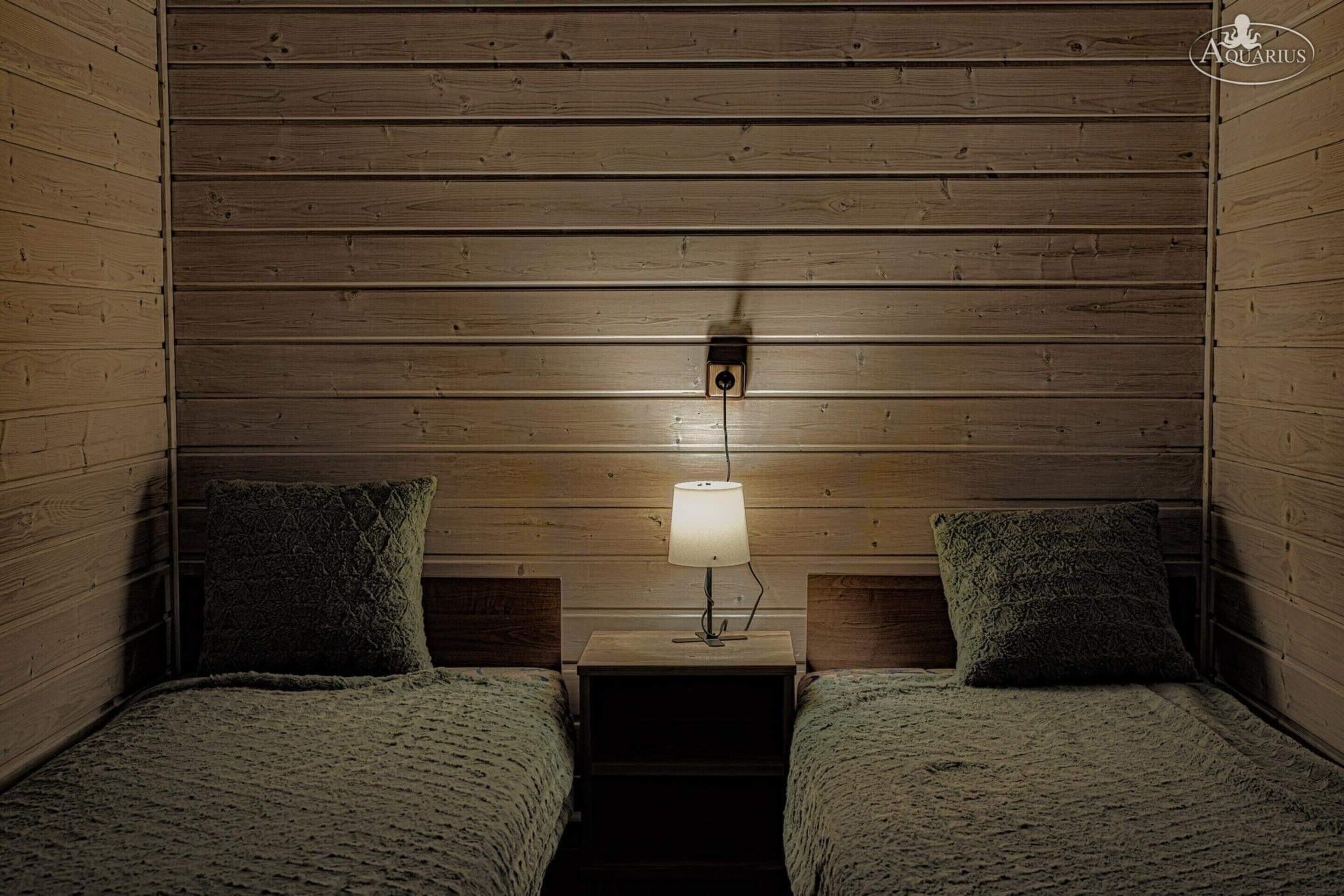 Ein gemütliches Zimmer mit Holzwandverkleidung, zwei Betten mit passenden Kissen sowie ein Nachttisch mit Lampe.