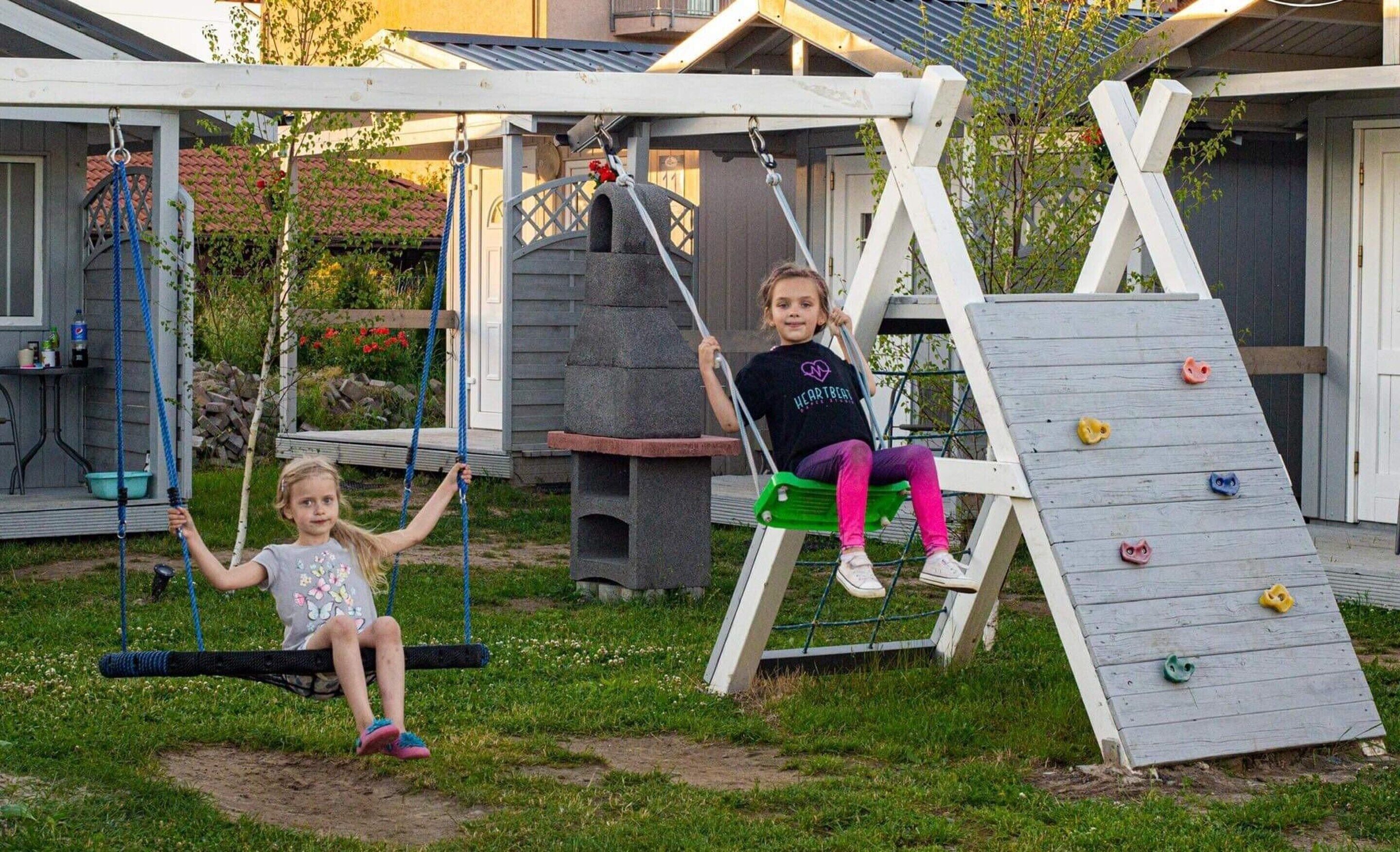 Two children on swings in a backyard with a climbing wall and a barbecue area.