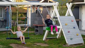 Two children on swings in a backyard with a climbing wall and a barbecue area.