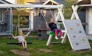 Two children on swings in a backyard with a climbing wall and a barbecue area.