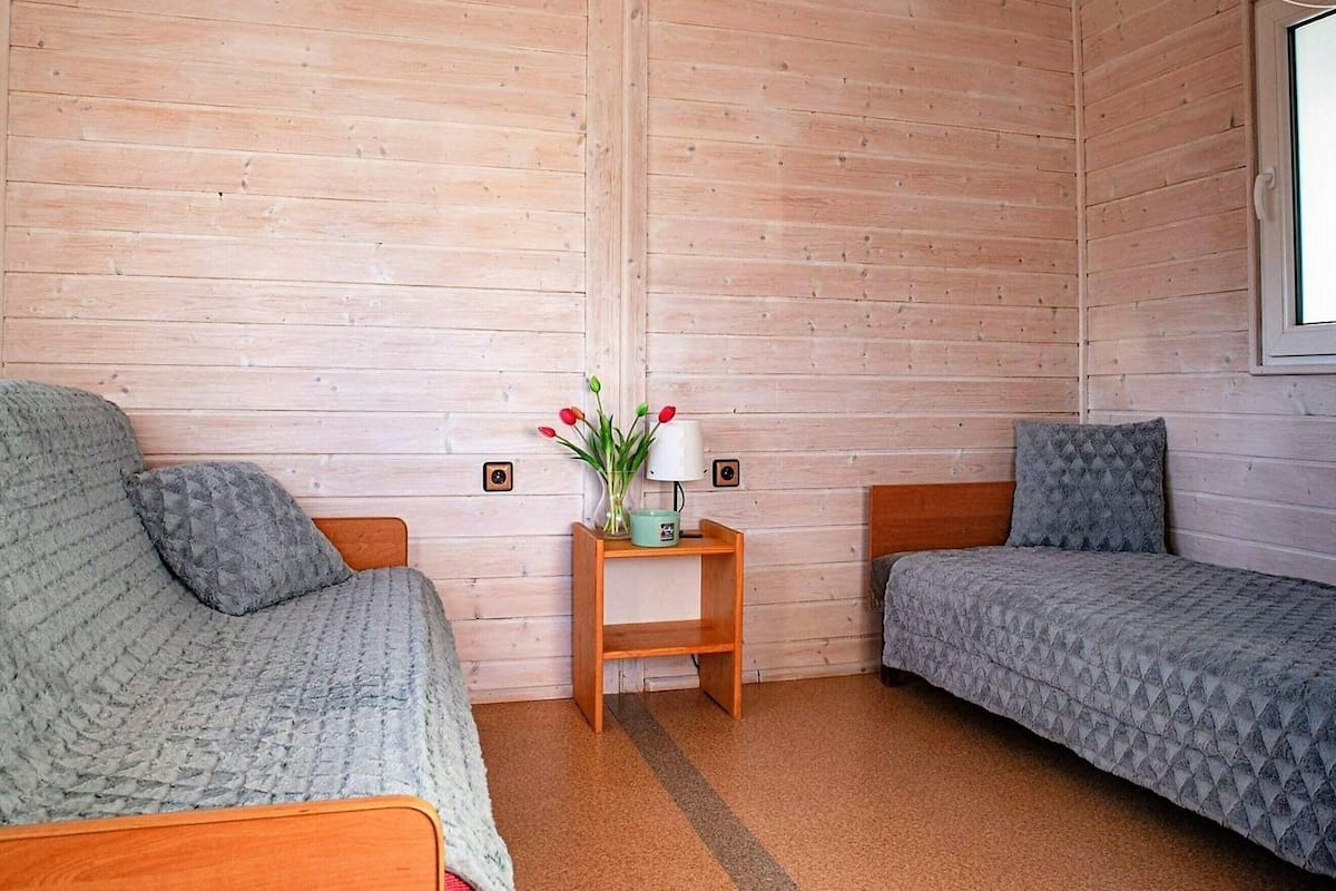 Una habitación pequeña y acogedora con dos camas individuales, una mesita de noche de madera con una lámpara y flores, y una ventana.