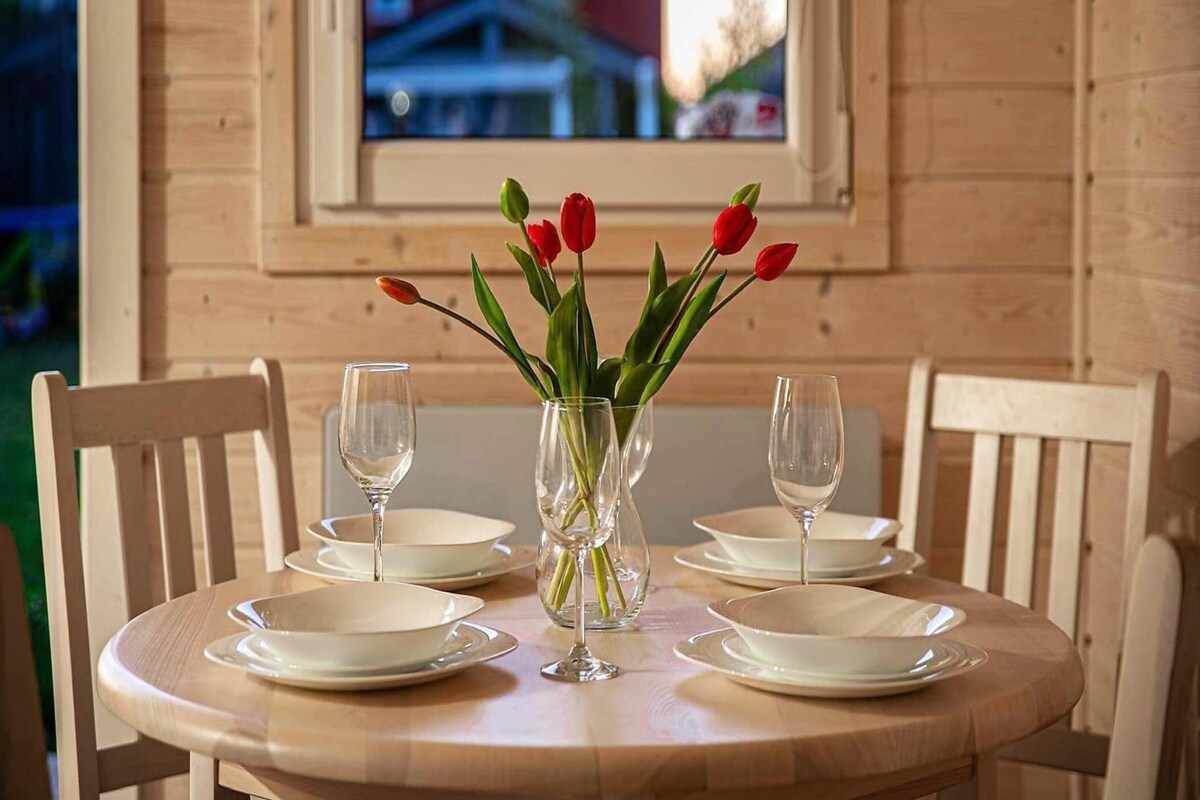 A wooden dining table with white plates and empty wine glasses, a vase with red tulips, and a window with a view of buildings.