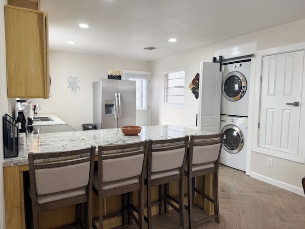 A kitchen with a marble countertop, wooden chairs, a built-in refrigerator, a washing machine, and a window with blinds.