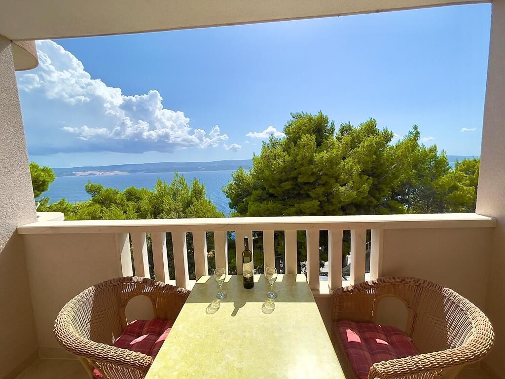 A balcony with wicker furniture, a table with wine glasses, and a view of the sea and trees.