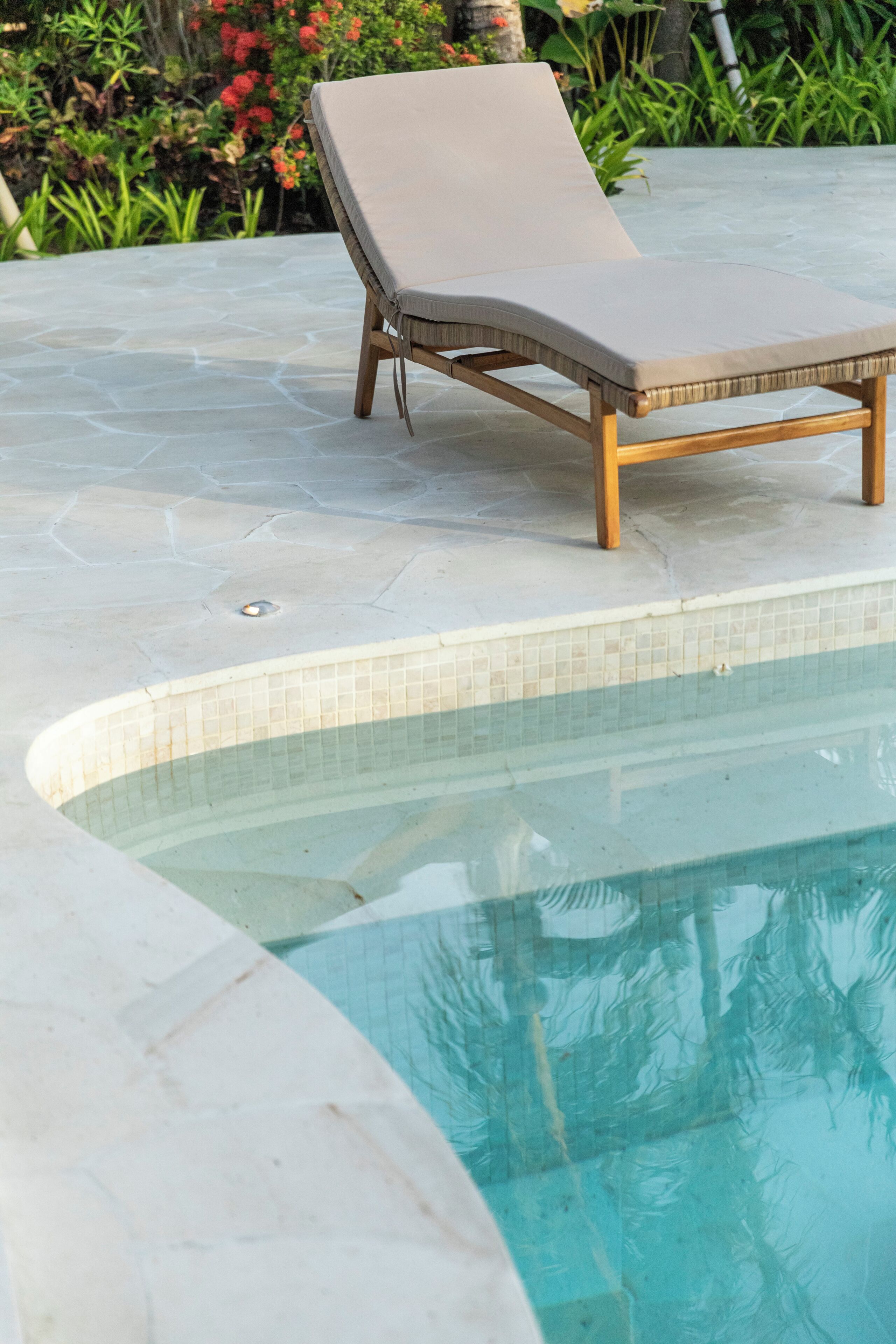 A poolside lounge chair with a cushioned seat and wooden legs, positioned on a stone patio next to a swimming pool.