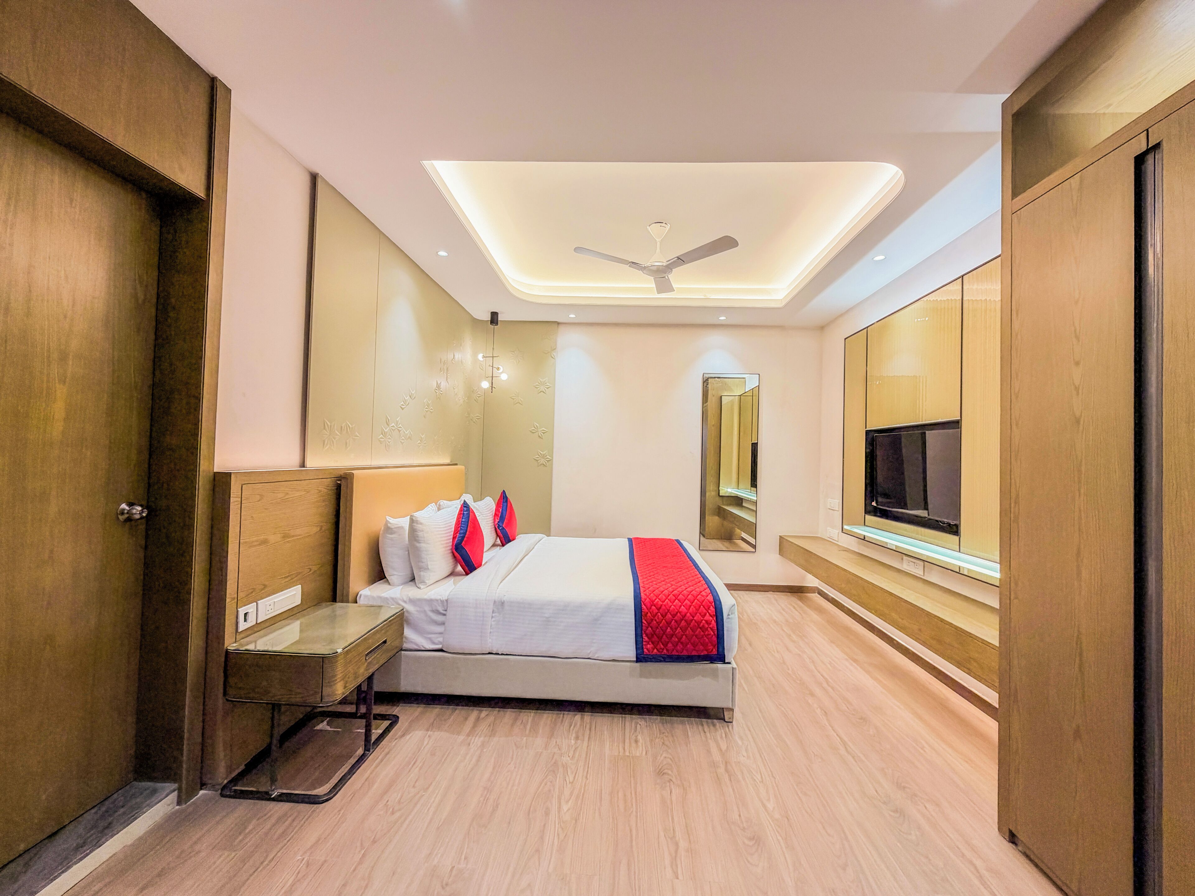 A modern hotel room with a bed, bedside table, mirror, TV, and ceiling fan.