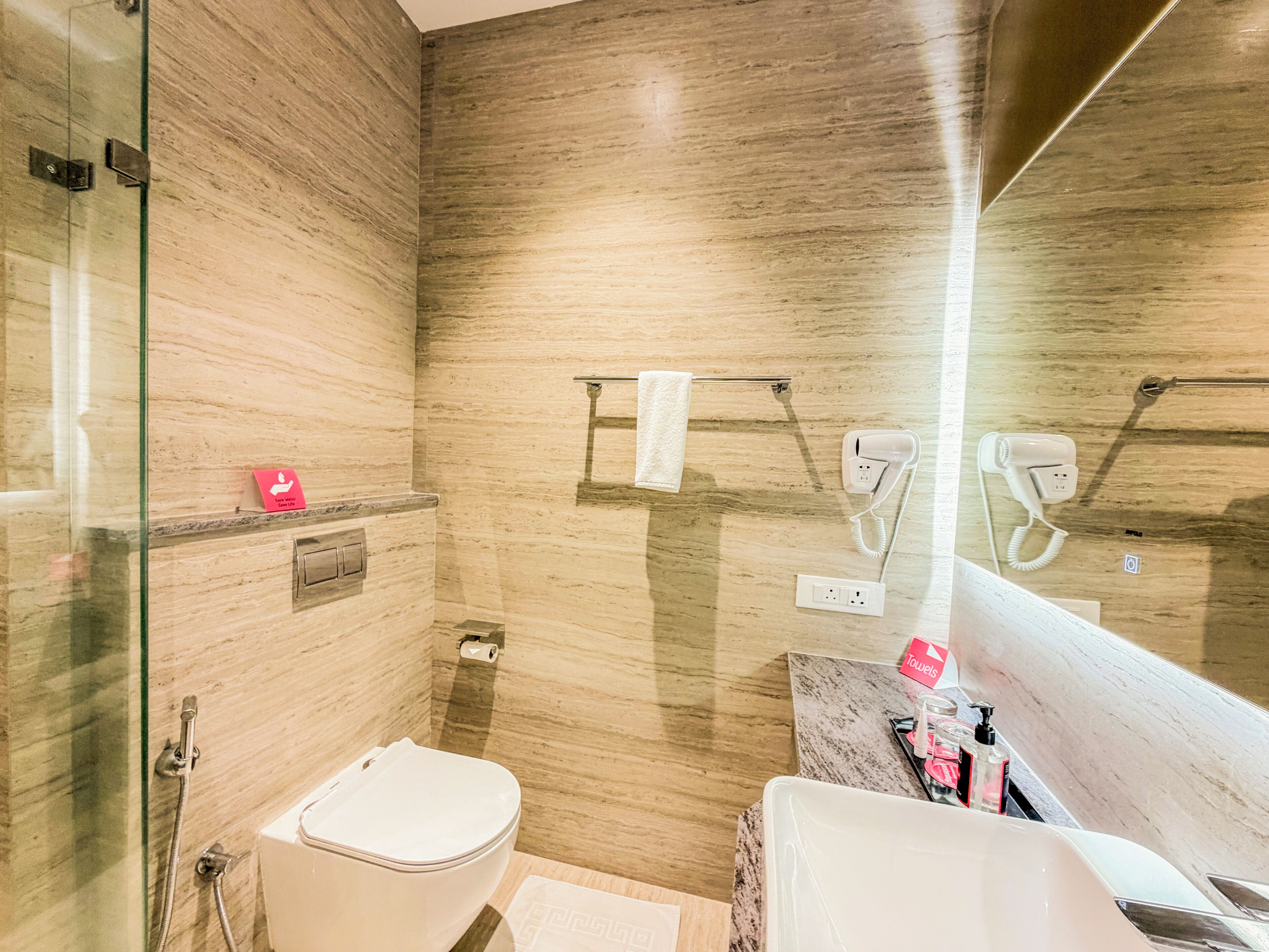 A modern bathroom with a marble wall, a glass shower enclosure, a toilet, a sink, and a towel rack with a hairdryer.