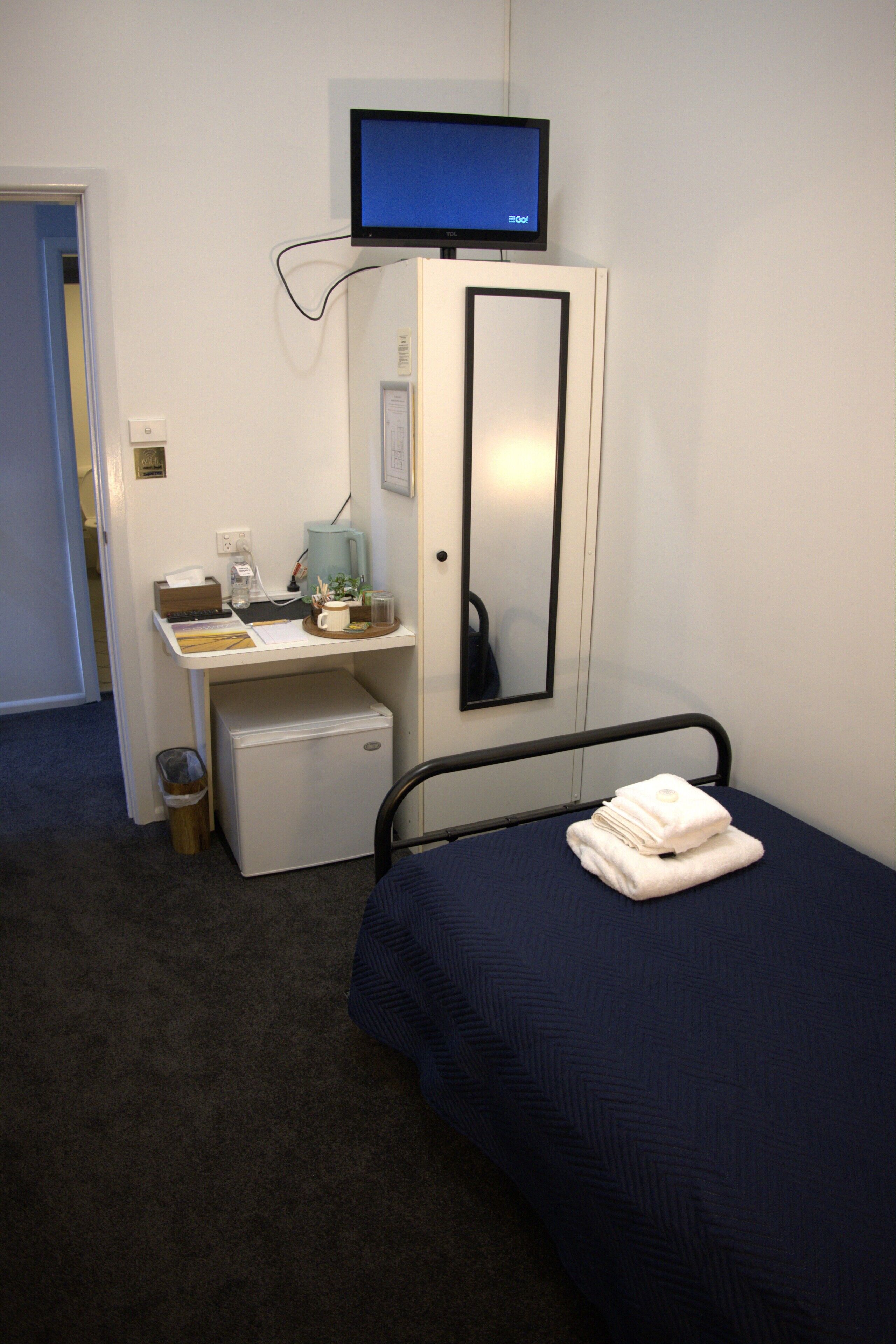 Habitación de hotel con cama, escritorio con computadora, refrigerador pequeño, televisor montado en la pared y un cubo de basura.
