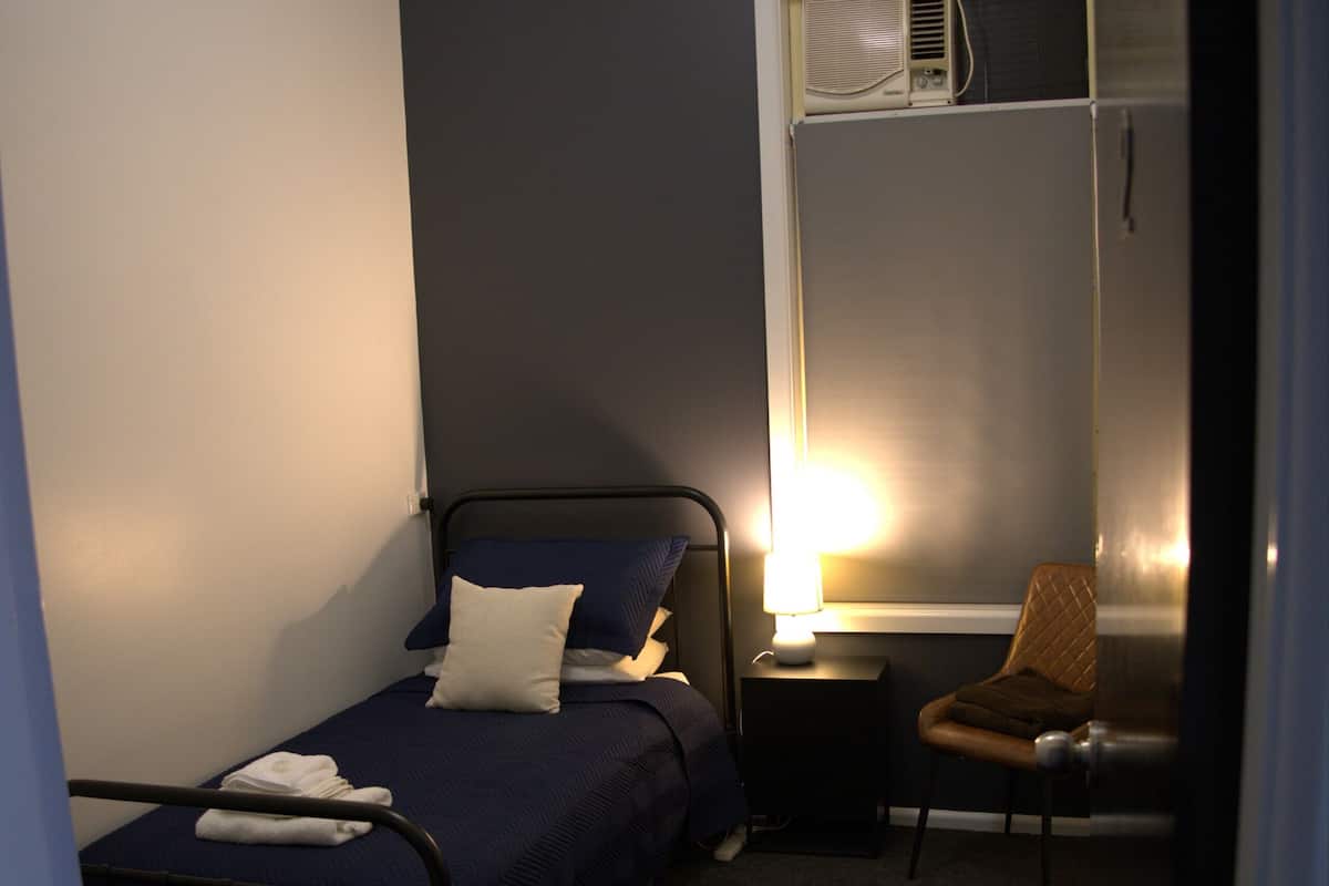 A small, dimly lit room with a single bed, a chair, a nightstand, and a window with an air conditioning unit.
