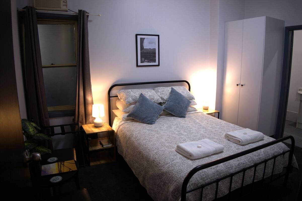 A neatly made bed with pillows and a blanket, a bedside table with a lamp, a wardrobe, and a framed picture on the wall.