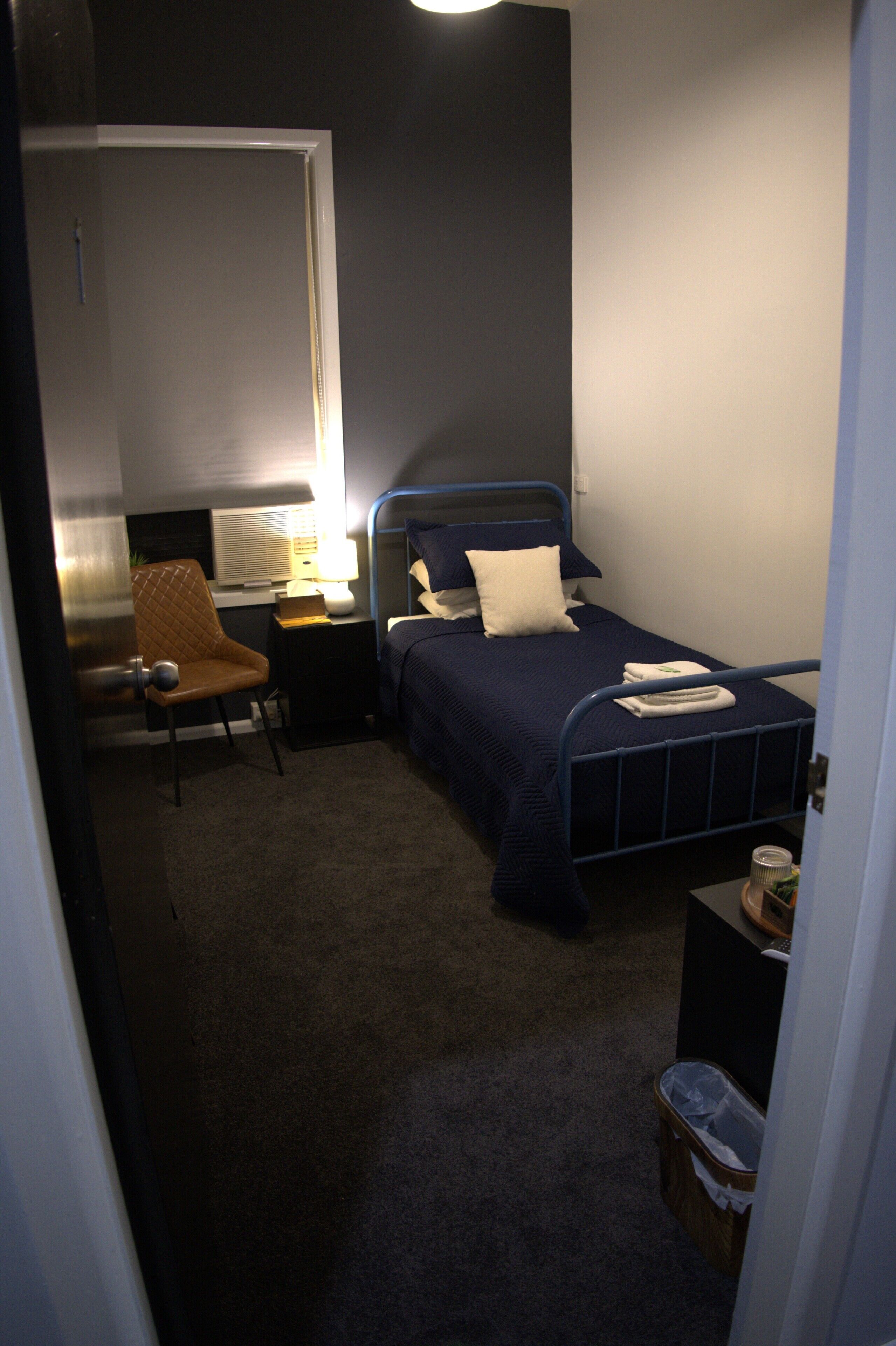 A bedroom with a single bed, a chair, a nightstand, and a small trash bin.