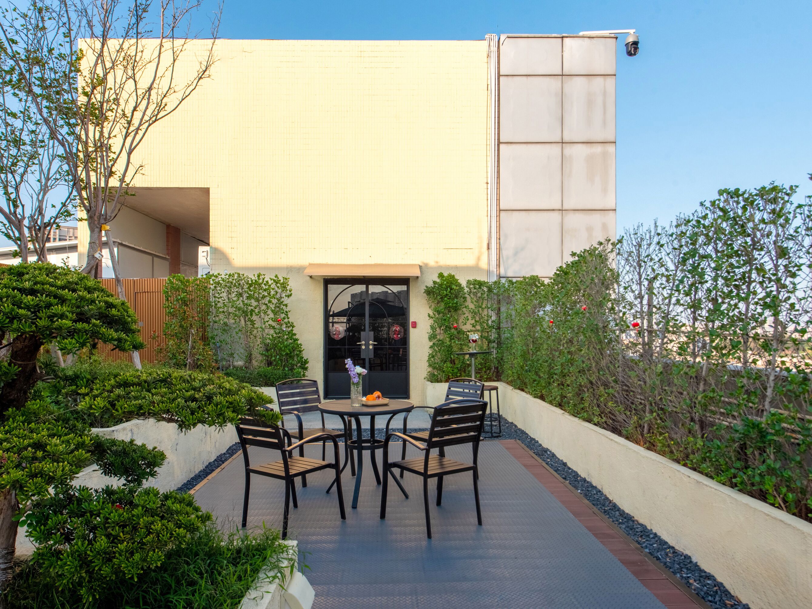 Un patio exterior con mesa y sillas, rodeado de follaje y la fachada de un edificio.