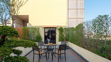 Un patio exterior con mesa y sillas, rodeado de follaje y la fachada de un edificio.