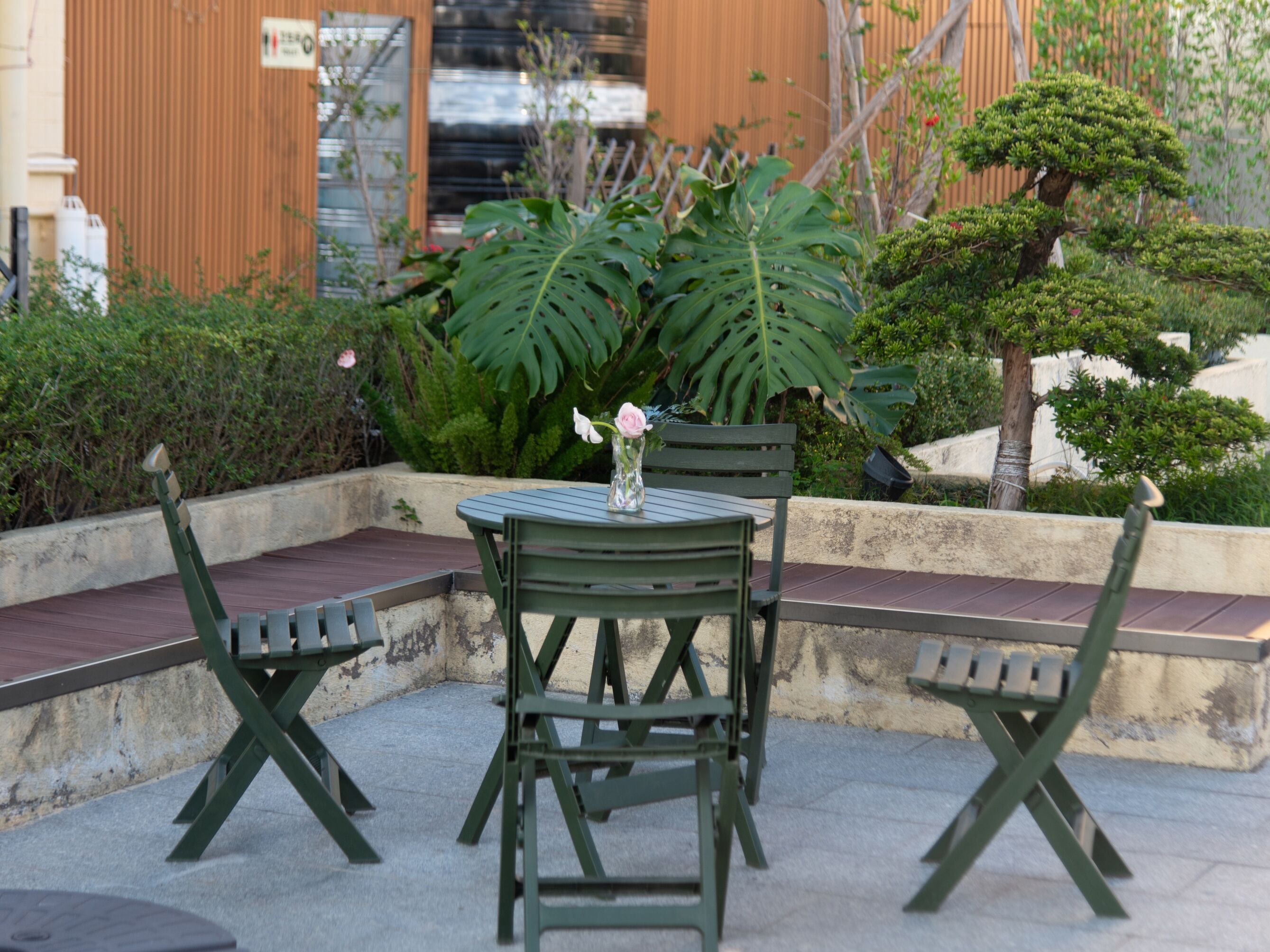 An outdoor seating area with a table and chairs, surrounded by greenery and a building in the background.