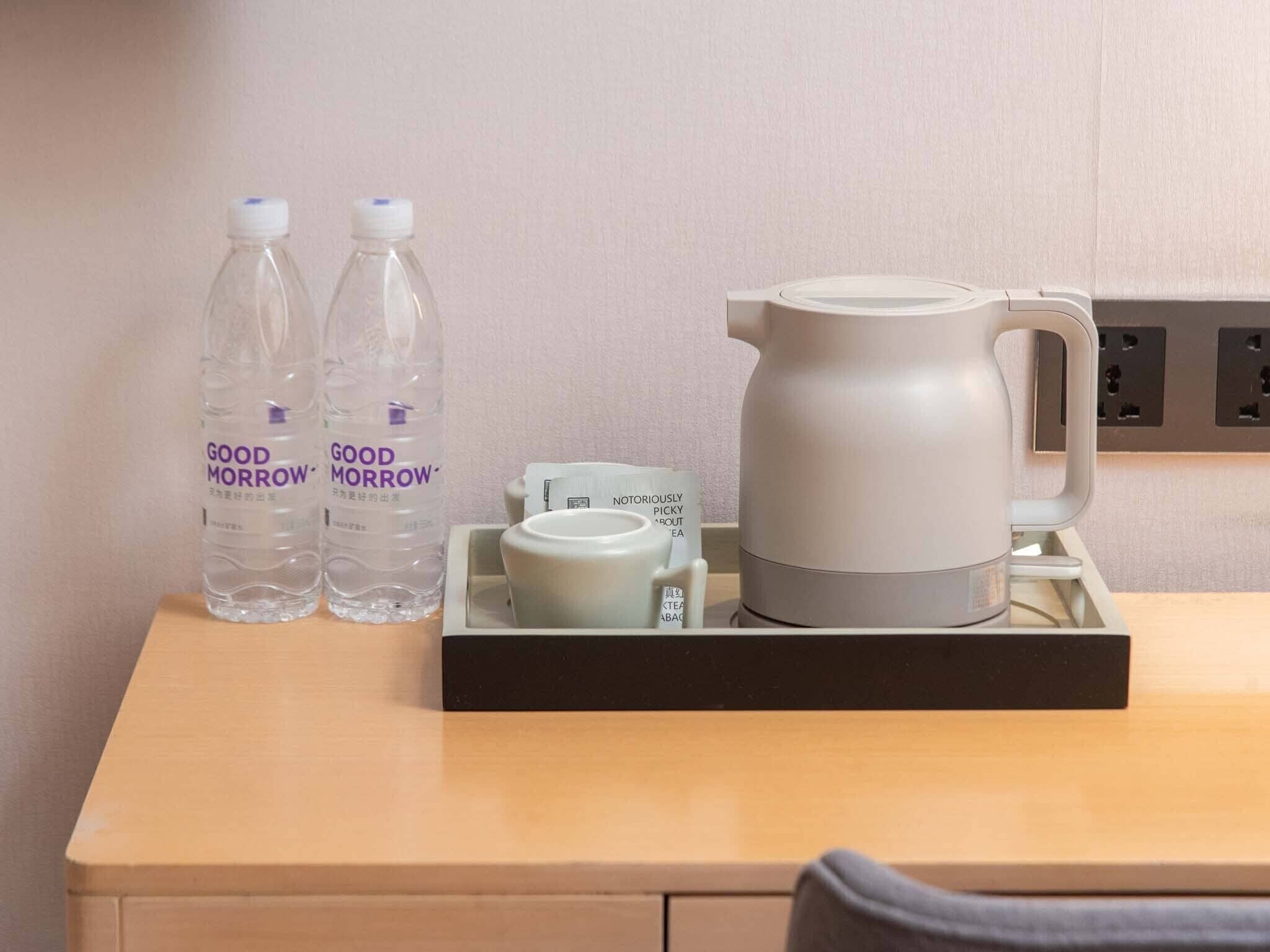A hotel room with a wooden table, two bottles of water, a white mug, and a gray electric kettle.