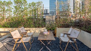 Un área de estar al aire libre con sillas plegables y una mesa, rodeada de follaje y un paisaje urbano de fondo.