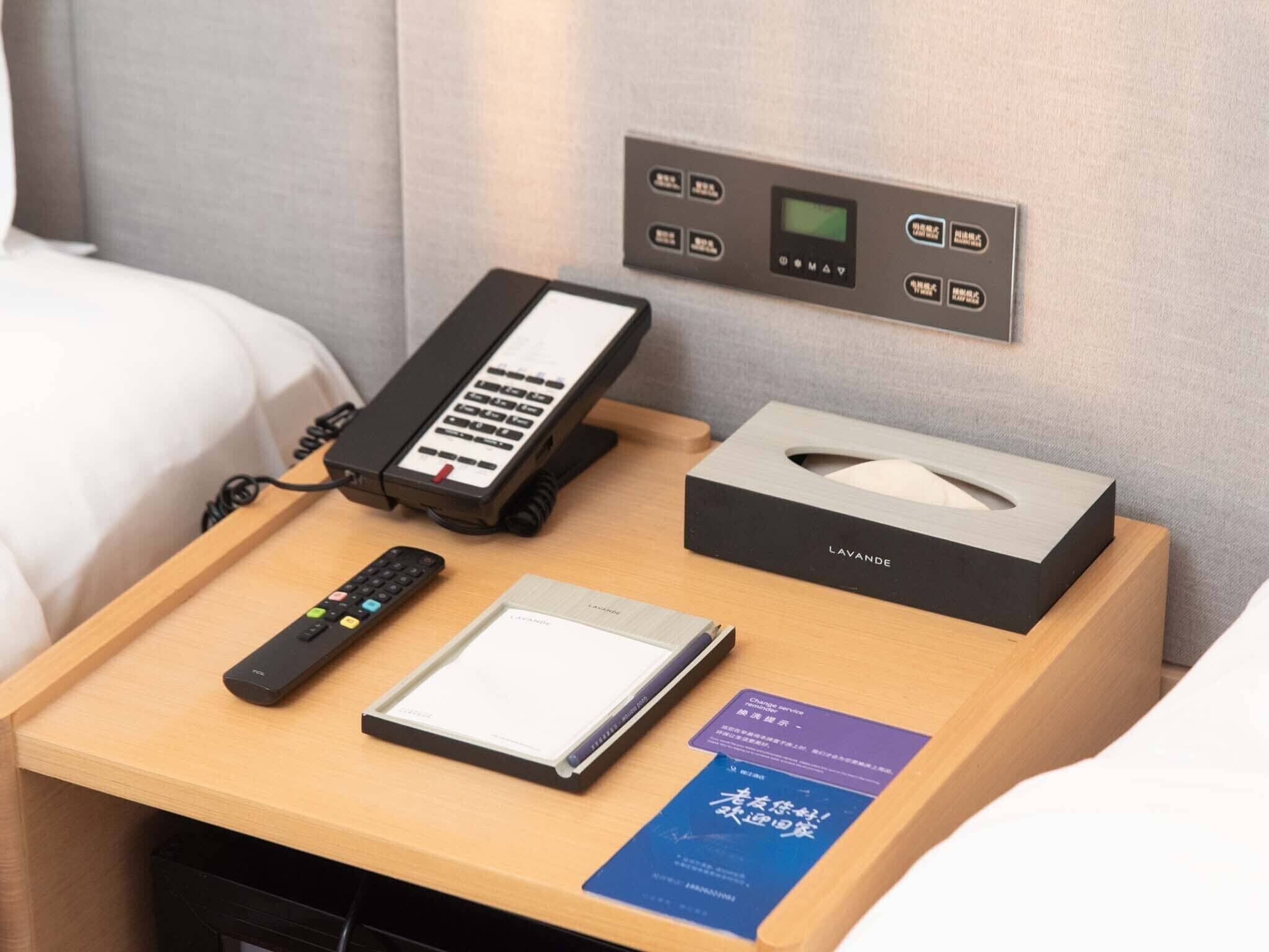 A hotel room with a bedside table featuring a telephone, remote control, tissue box, and a booklet.