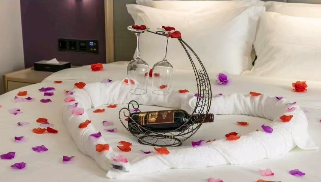 Un lit recouvert de pétales de rose, deux verres à vin posés sur un porte-verres métallique et une bouteille de vin.