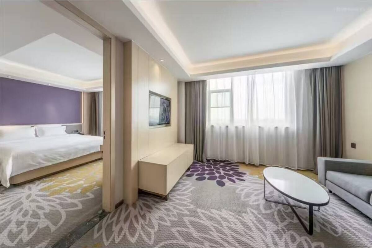 Una habitación de hotel con cama, sofá, una mesita y un amplio ventanal con cortinas.