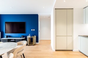 A modern living room with a blue accent wall, a flat-screen TV, a dining area with a marble table, and a kitchen with white cabinets. - Stylish 2-Bed by The O2 with Balcony (London)