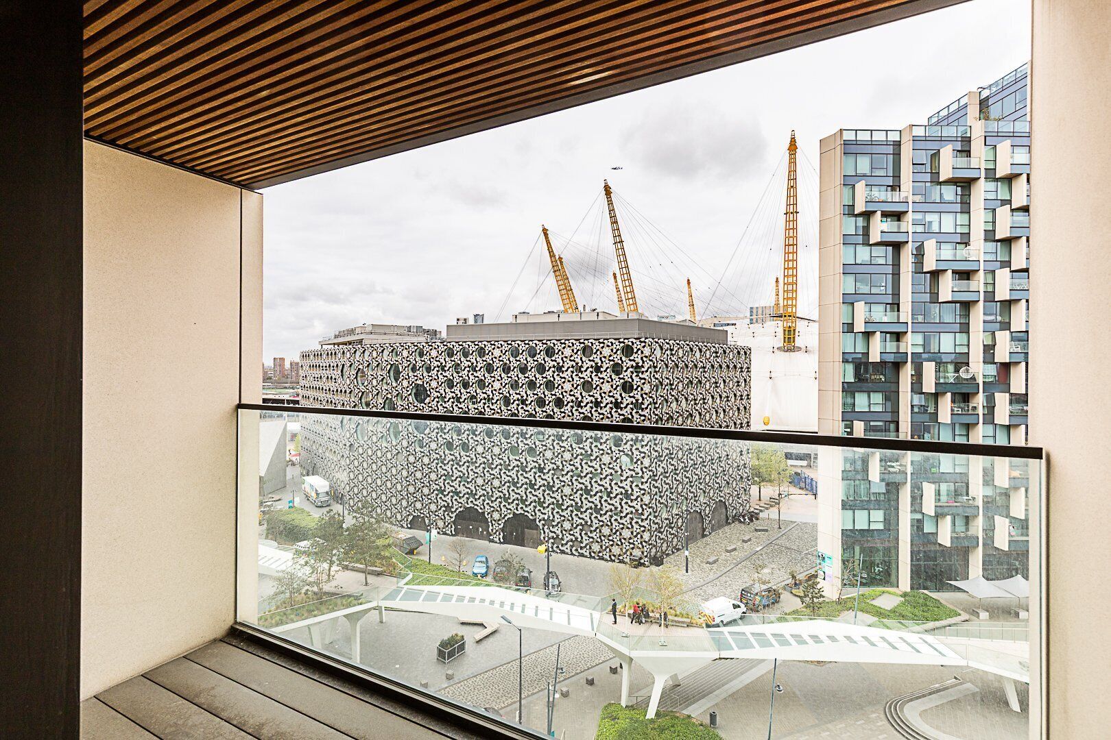Ein modernes Gebäude mit einer markanten Fassade, einem Balkon mit Holzboden und Blick auf eine Baustelle mit Kränen.