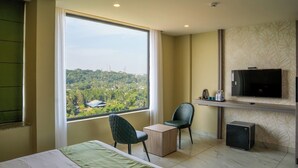 Une chambre d’hôtel avec un lit, deux chaises, une télévision, une petite table et une grande fenêtre donnant sur un paysage verdoyant.