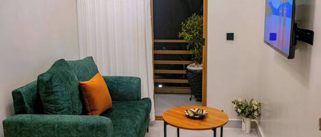 A living room with a green sofa, a small wooden table, a TV mounted on the wall, and a potted plant.