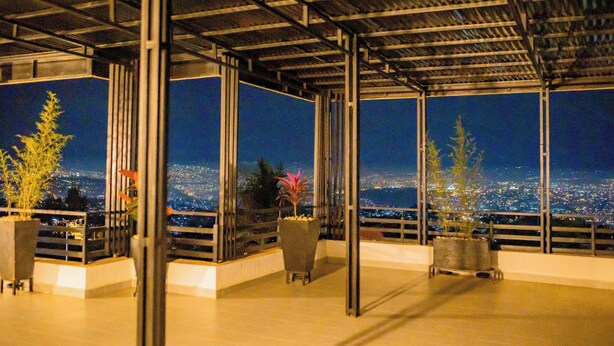 Un espacio al aire libre techado con vistas a un paisaje urbano nocturno, con columnas metálicas, plantas en macetas y una barandilla.