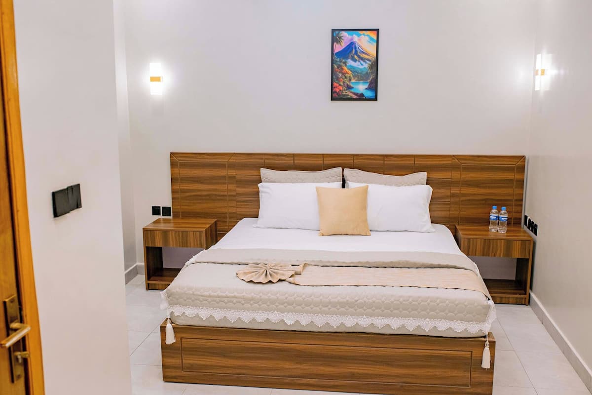 Une chambre à coucher bien rangée, avec une tête de lit en bois, un lit recouvert de draps blancs, deux tables de chevet avec des bouteilles d’eau et un tableau accroché au mur.