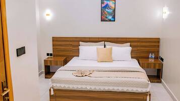 Un dormitorio ordenado con cabecera de madera, cama con ropa blanca, dos mesitas de noche con botellas de agua y un cuadro en la pared.