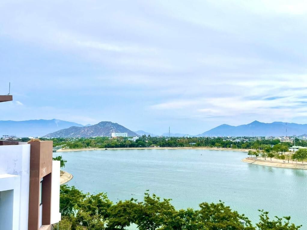 A view of a lake with mountains in the background, a building with a flat roof in the foreground, and a clear sky.