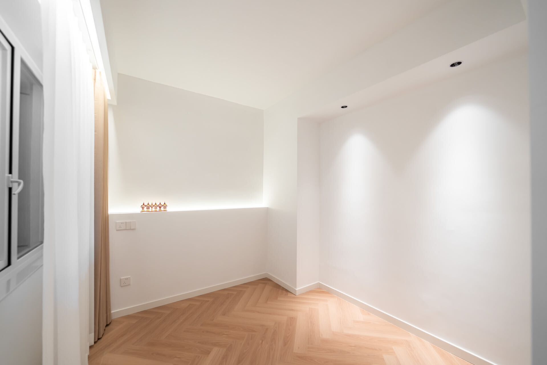 Una habitación con piso de madera en patrón de espiga, paredes blancas y una ventana pequeña con una lámpara.