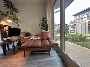 A modern living room with a brown leather sofa, a desk with a computer, a chair, and large windows offering a view of a building and greenery outside. - Serene 1-bedroom townhome with AC near ATL Airport (College Park)