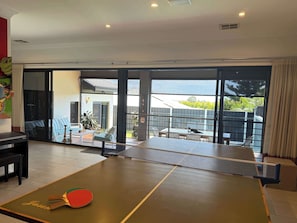 Une table de ping-pong se trouve dans une pièce dotée d’une grande porte-fenêtre coulissante qui donne sur un espace extérieur offrant une vue sur une maison et sur la verdure.