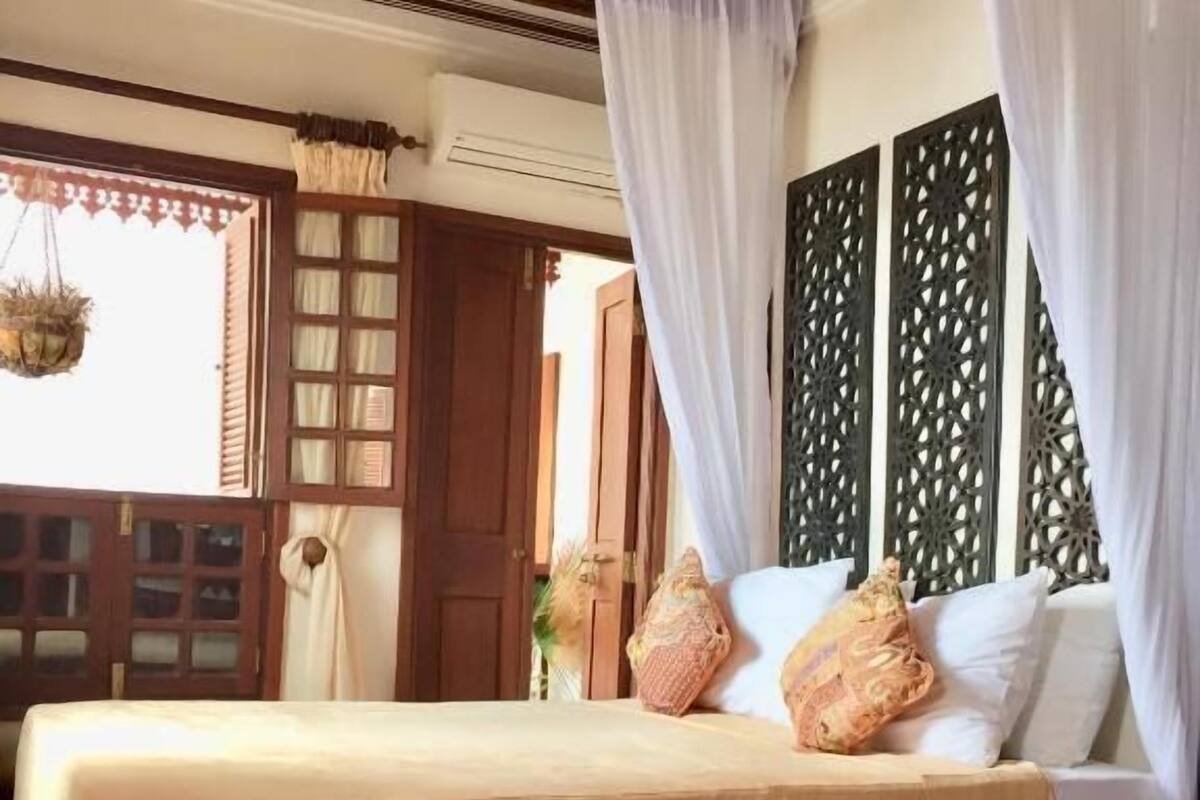 A bed with a canopy, a wooden headboard, a patterned floor mat, and a decorative hanging.