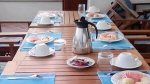 Una mesa de comedor dispuesta con platos, cubiertos y una cafetera, con un sofá y sillas al fondo.
