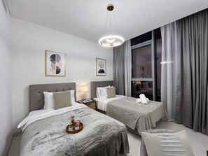 Ein Hotelzimmer mit zwei Betten, einem Nachttisch, einer Lampe, einem Fenster mit Vorhängen und einem Kunstwerk an der Wand.