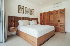 Un dormitorio con estructura de cama de madera, una mesita de noche con lámpara, un armario y tres láminas botánicas enmarcadas en la pared.