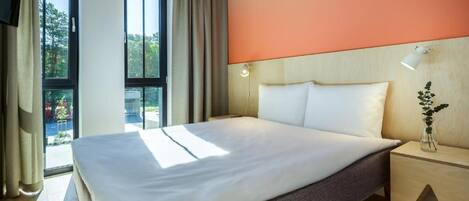 A hotel room with a large bed, a nightstand with a lamp, a view of the outdoors, and a window with curtains.
