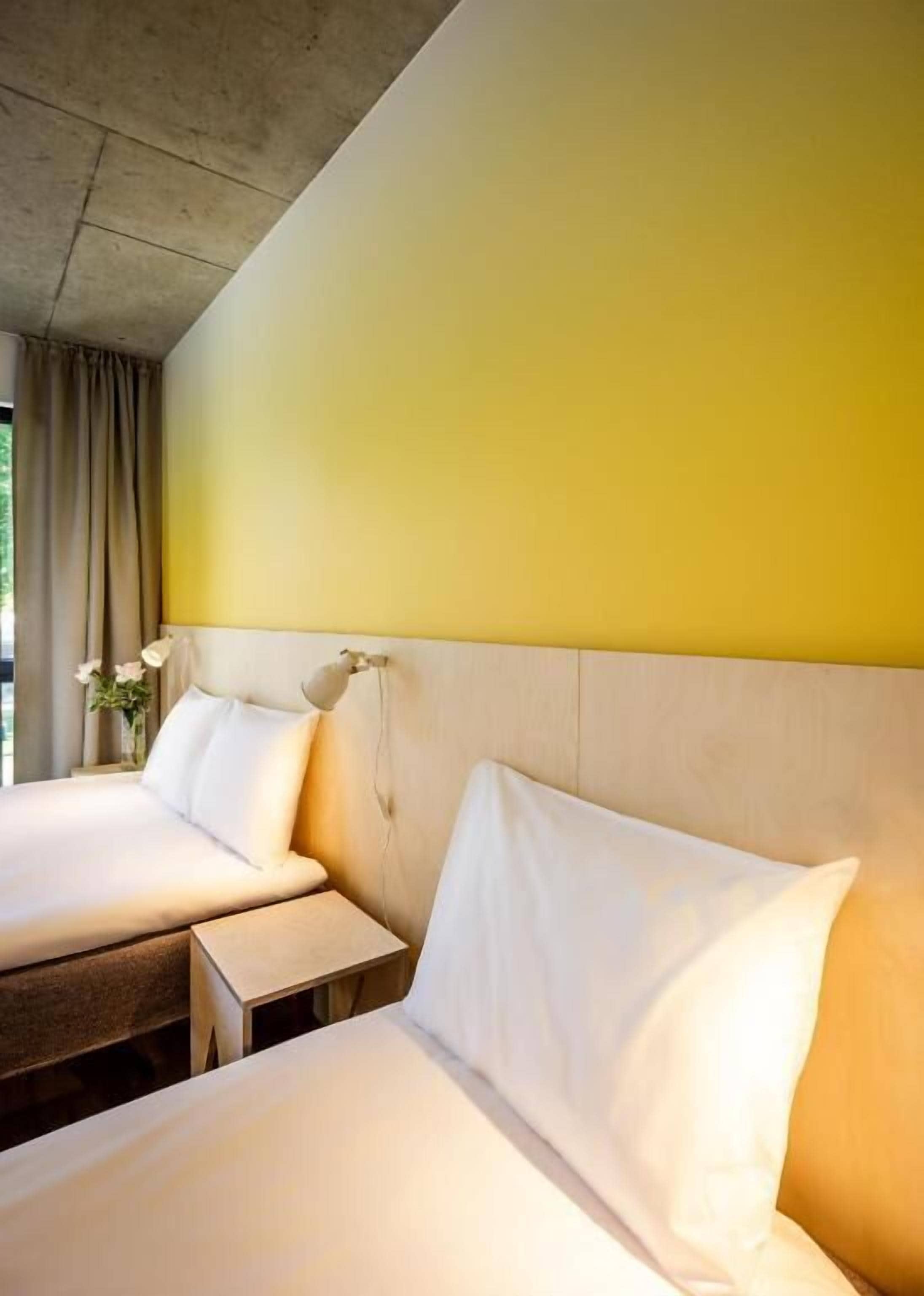 A hotel room with two beds, a bedside table, a lamp, and a window with curtains.