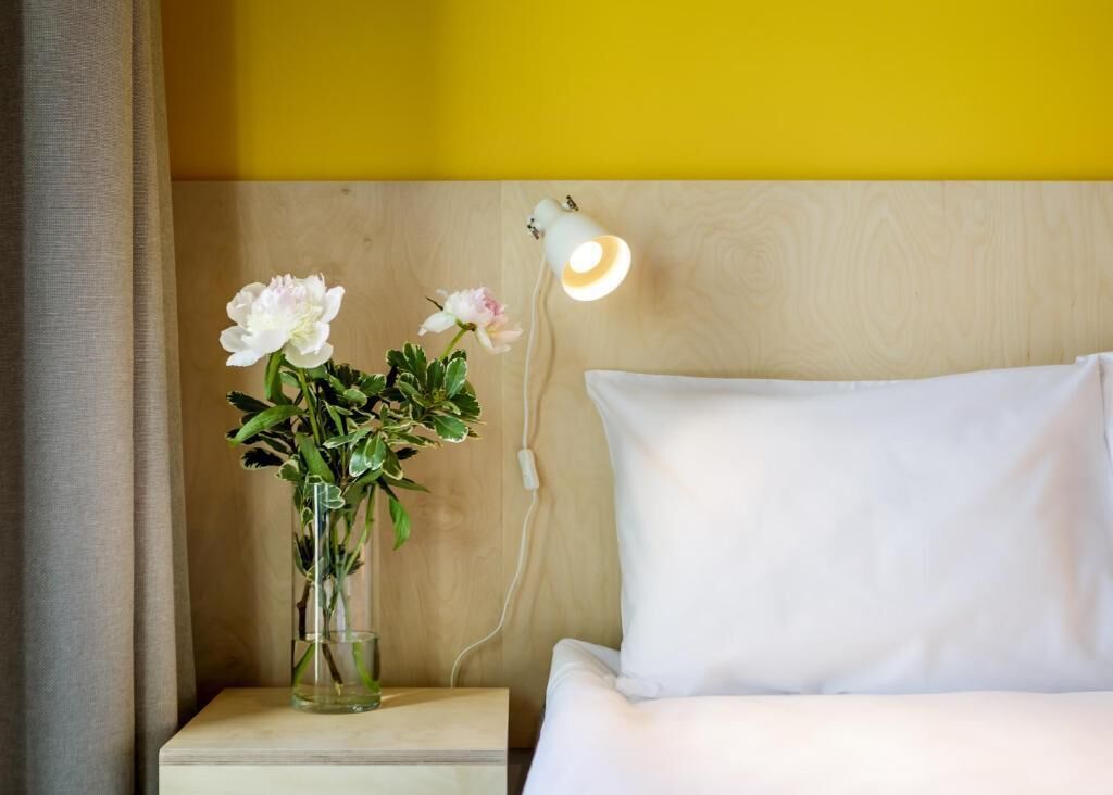 A bed with white bedding, a bedside lamp, and a clear vase with flowers on a wooden nightstand.