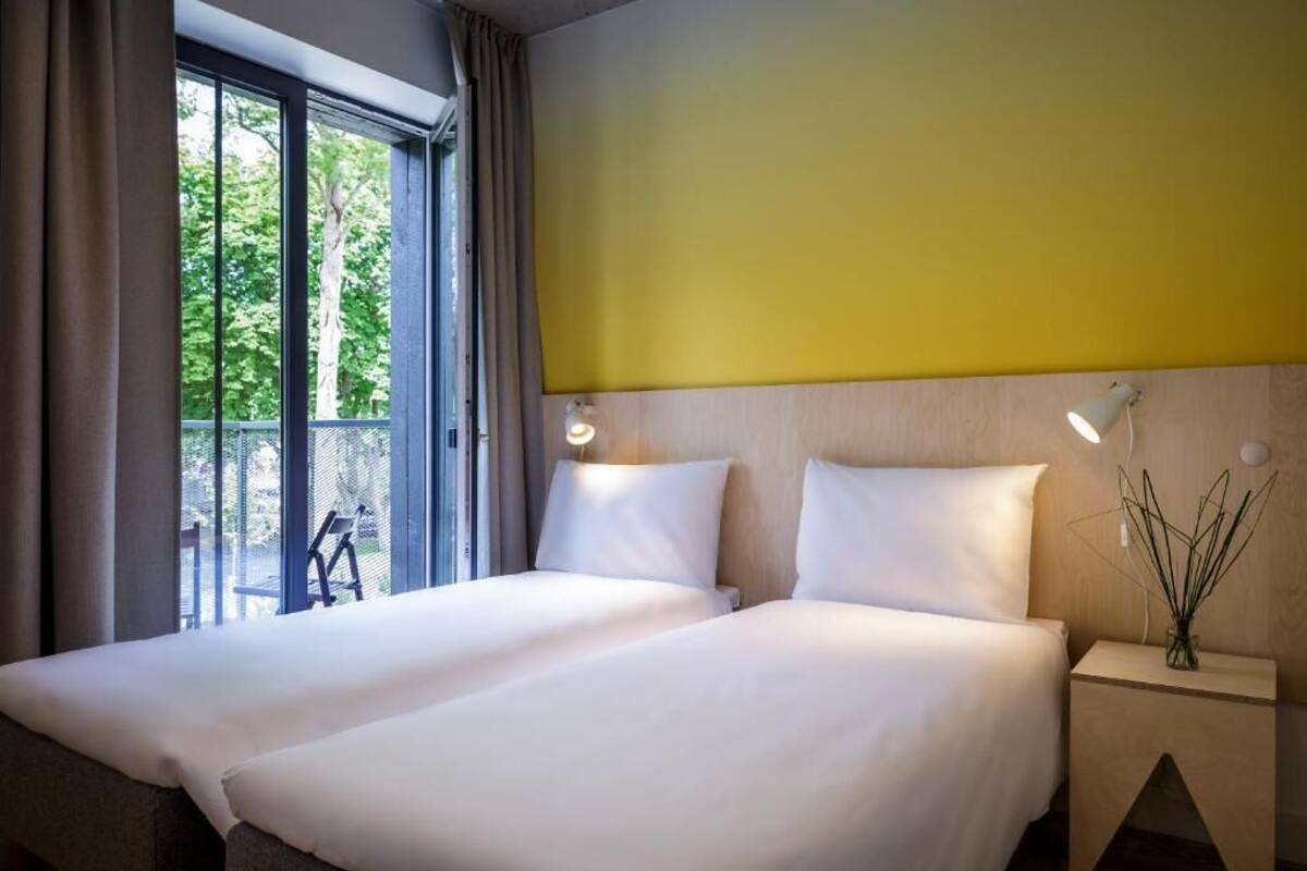 Habitación de hotel con dos camas, un ventanal con cortinas, cabecera de madera y una mesita de noche con lámpara y florero decorativo.