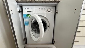 Een compacte, witte wasmachine met voorlader in een technische ruimte, met een granieten aanrechtblad en een houten vloer.