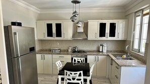 Een moderne keuken met witte kasten, een roestvrijstalen koelkast, een eettafel met stoelen en een raam met jaloezieën.