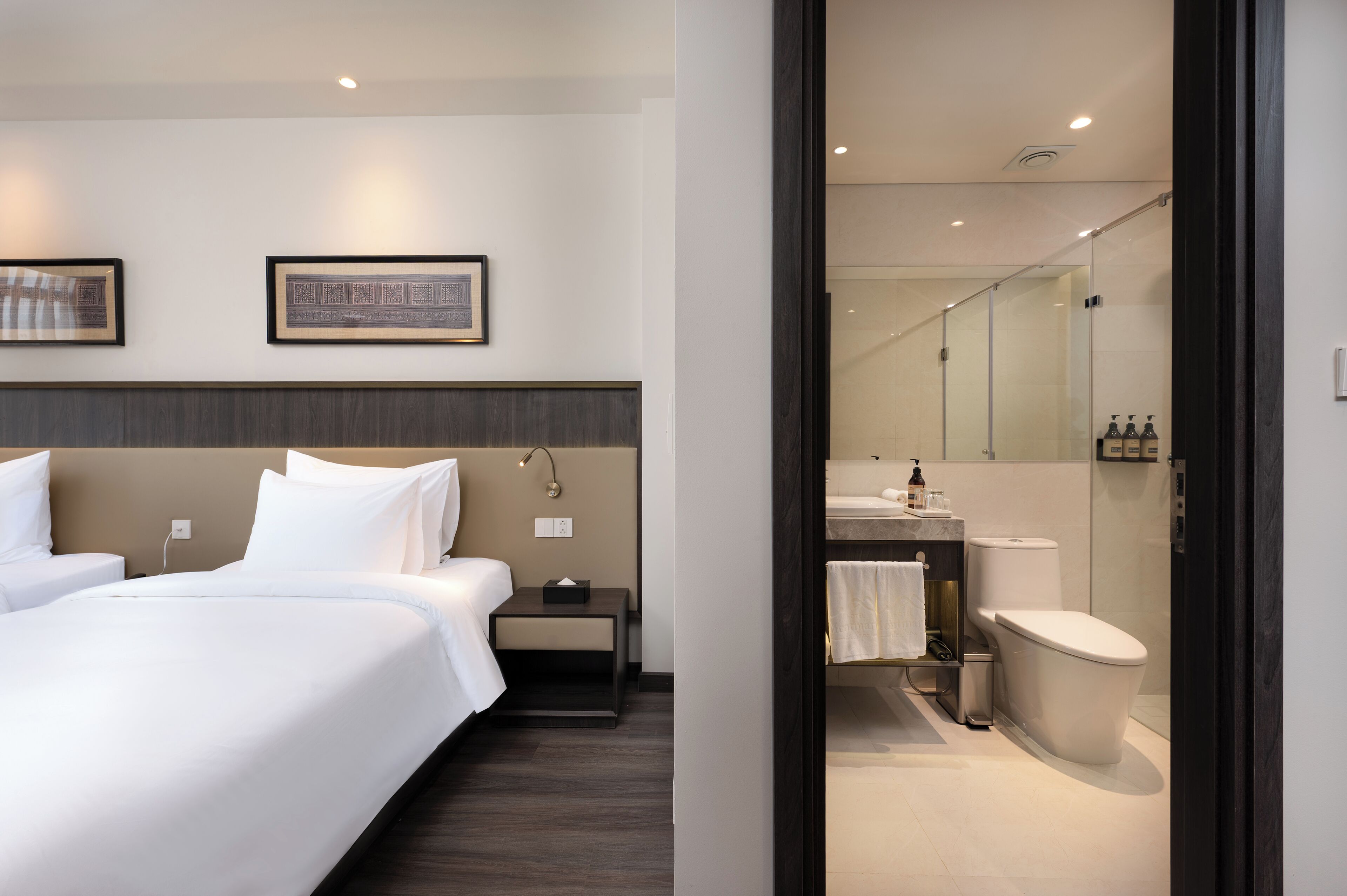 A hotel room with two beds, a nightstand, a wall-mounted lamp, and a framed picture.