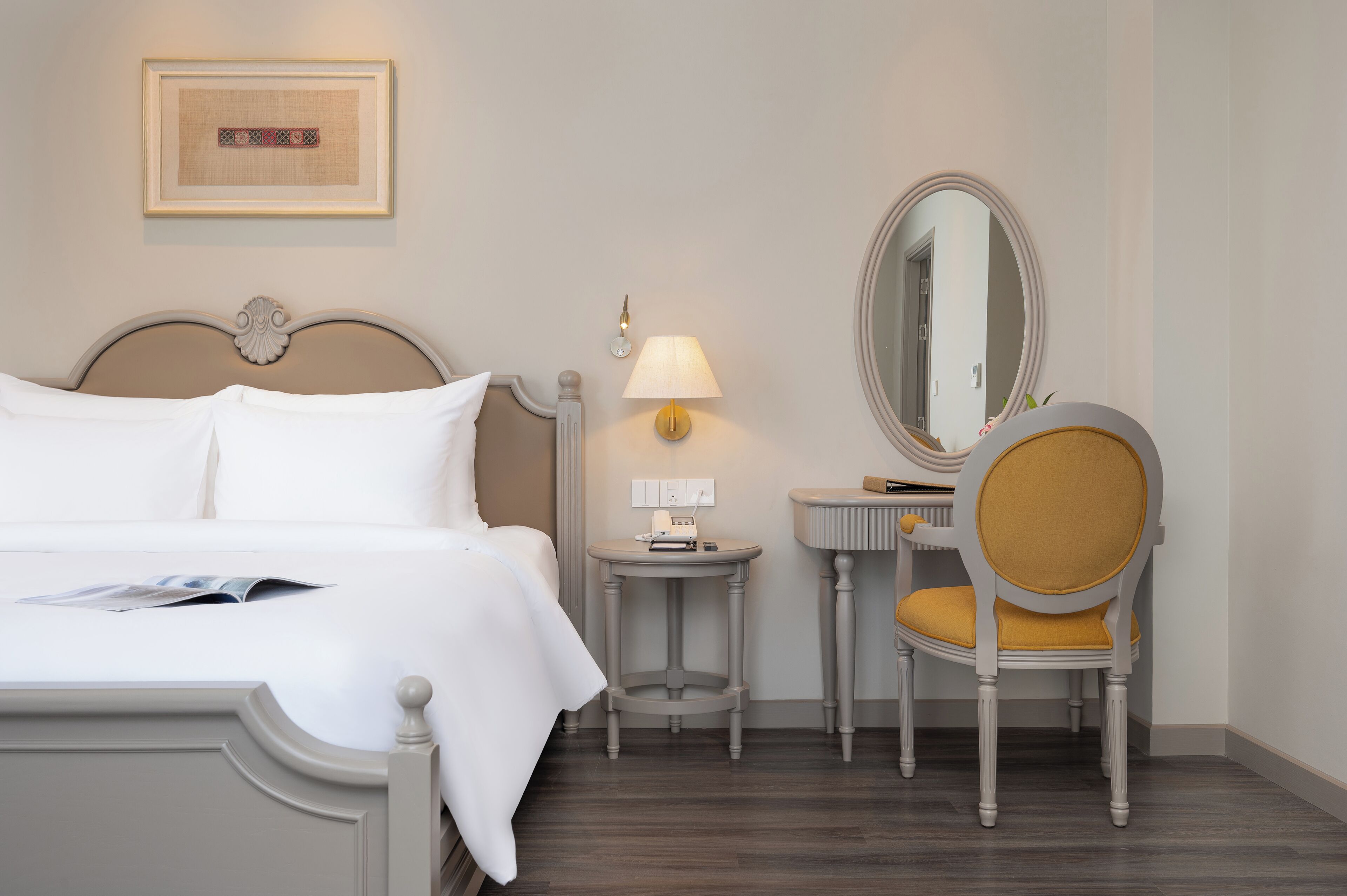 A hotel room with a bed, bedside table, lamp, mirror, and a chair with a cushion.