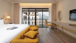 A hotel room with a large bed, a sofa, a desk, a chair, a TV, and a balcony with a view of buildings and mountains. - Hotel Montmartre de Sapa (Sa Pa)