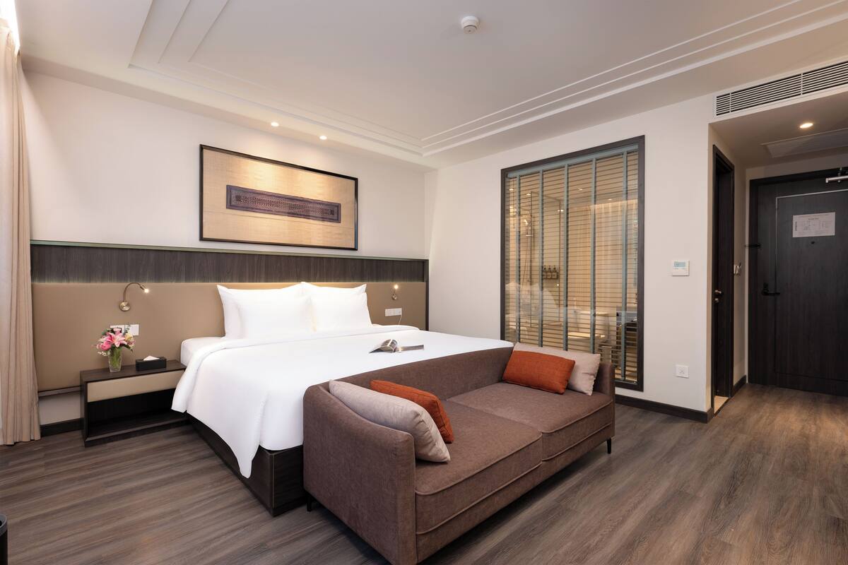 Habitación de hotel con cama, sofá, mesita de noche, una obra de arte en la pared, ventana con persianas y puerta.